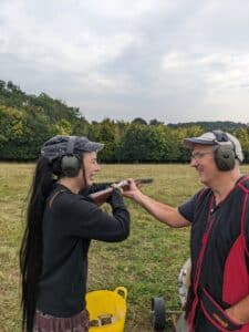 Two people in protective ear gear stand on a grassy field, holding a shotgun together, with one instructing the other. Trees are visible in the background.